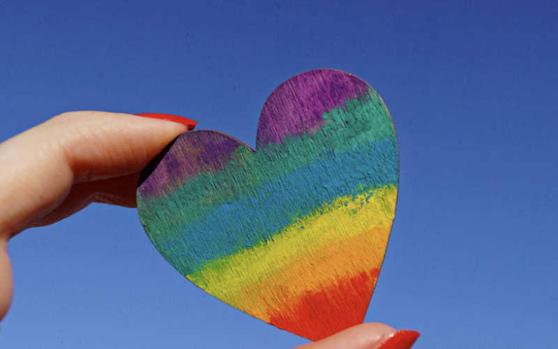 a hand with fingernails painted red holds a tiny rainbow heart up in the air